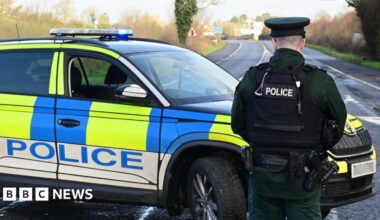 A police officer is standing on a road with his back towards the camera. A blue, yellow and white police car is in front of him. No traffic is on the road.