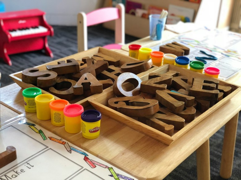 The middle of a wooden table is filled with large wooden blocks styled into letters while on the sides are colourful containers of play dough. In the background of the brightly lit daycare room, a bright red piano and a bookshelf are visible.