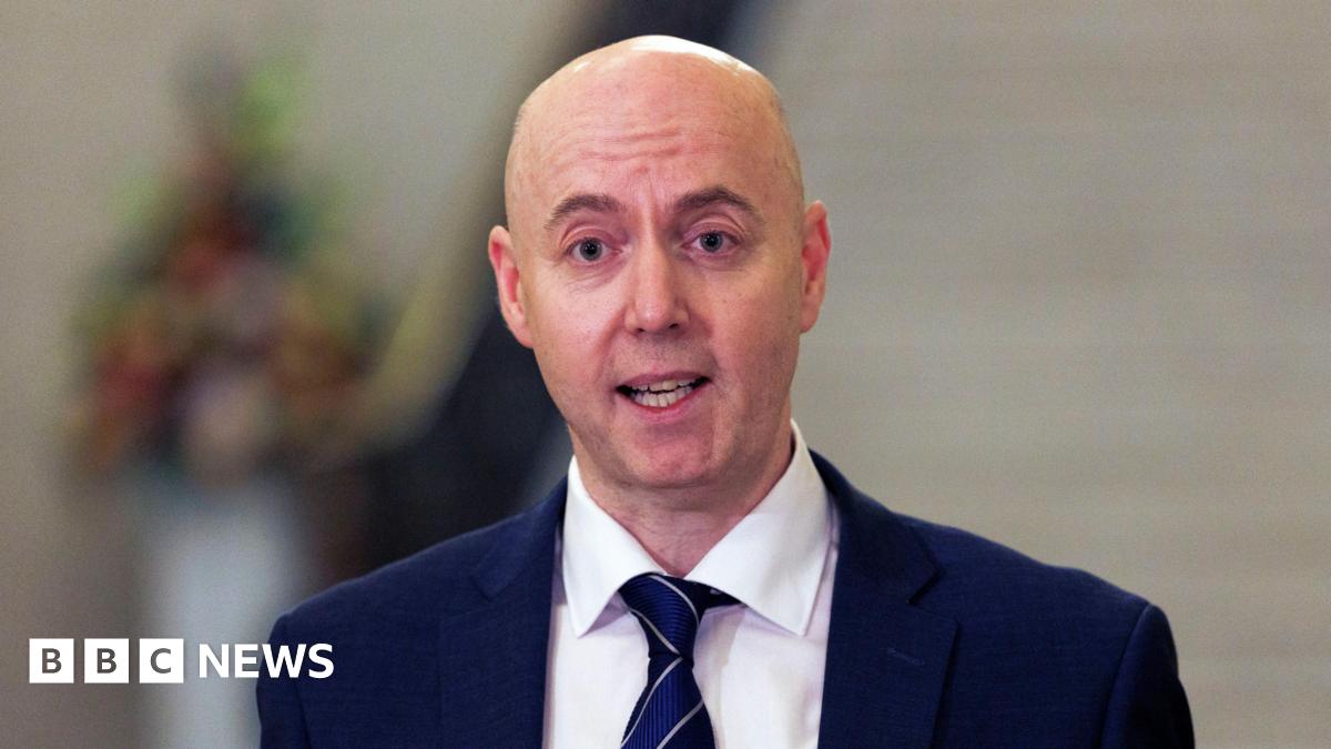 Jon Burrows is wearing a white shirt with navy blazer and tie. The background is blurry.