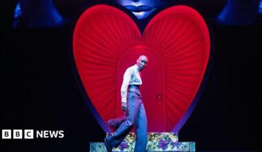 Cynthia Erivo stands in front of a heart on stage in a suit while a visual projection of her face is above it.