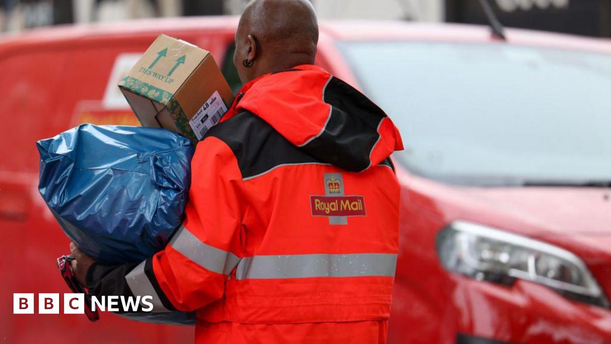 A Royal Mail postman holding two large parcels. He is standing in front of a red Royal Mail van