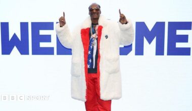 Snoop Dogg wears a fur coat over a red tracksuit, fingers pointed upwards while standing in front of a large sign saying 'Welcome' at the Winter Olympics