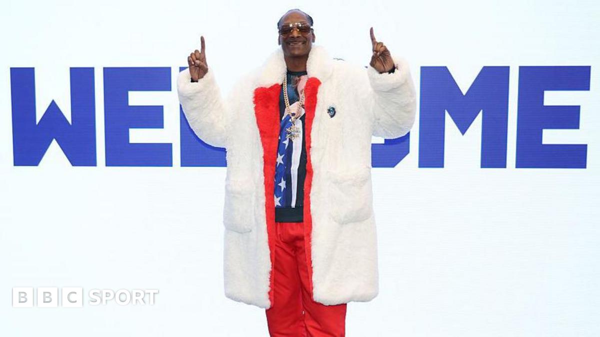 Snoop Dogg wears a fur coat over a red tracksuit, fingers pointed upwards while standing in front of a large sign saying 'Welcome' at the Winter Olympics