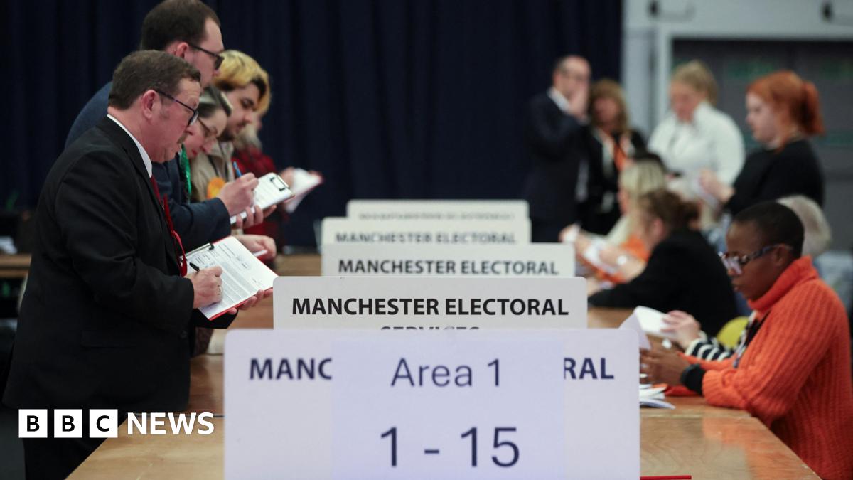 Votes are counted for the Gorton and Denton by-election at the Manchester Central Convention Complex. A row of people standing and wearing party rosettes stands to the left of a series of tables while vote counters sit on the other side.