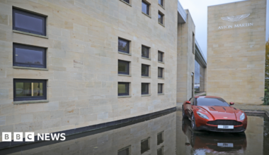 A red Aston Martin car is displayed outside the company's manufacturing site in Warwickshire. The building has white bricks and Aston Martin plus its logo displayed on it in silver.