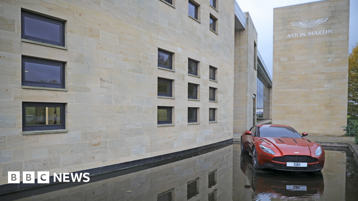 A red Aston Martin car is displayed outside the company's manufacturing site in Warwickshire. The building has white bricks and Aston Martin plus its logo displayed on it in silver.