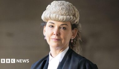 Dorothy Bain, who has long dark hair tied back under a court wig, looks straight at the camera.