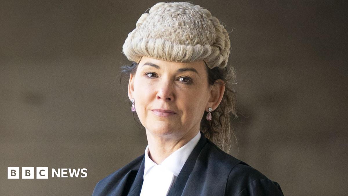 Dorothy Bain, who has long dark hair tied back under a court wig, looks straight at the camera.