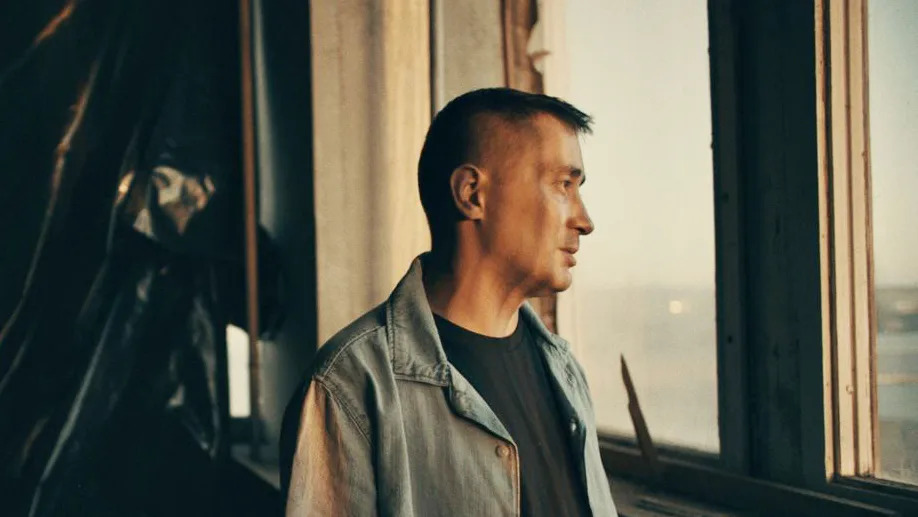 Ilya, wearing a blue denim jacket, looks out of the window in a warehouse in an undisclosed location.