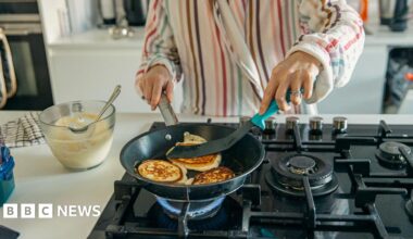 A woman dressed in striped pyjamas cooks pancakes over a gas stovetop. A glass bowl of pancake batter is on the bench beside her