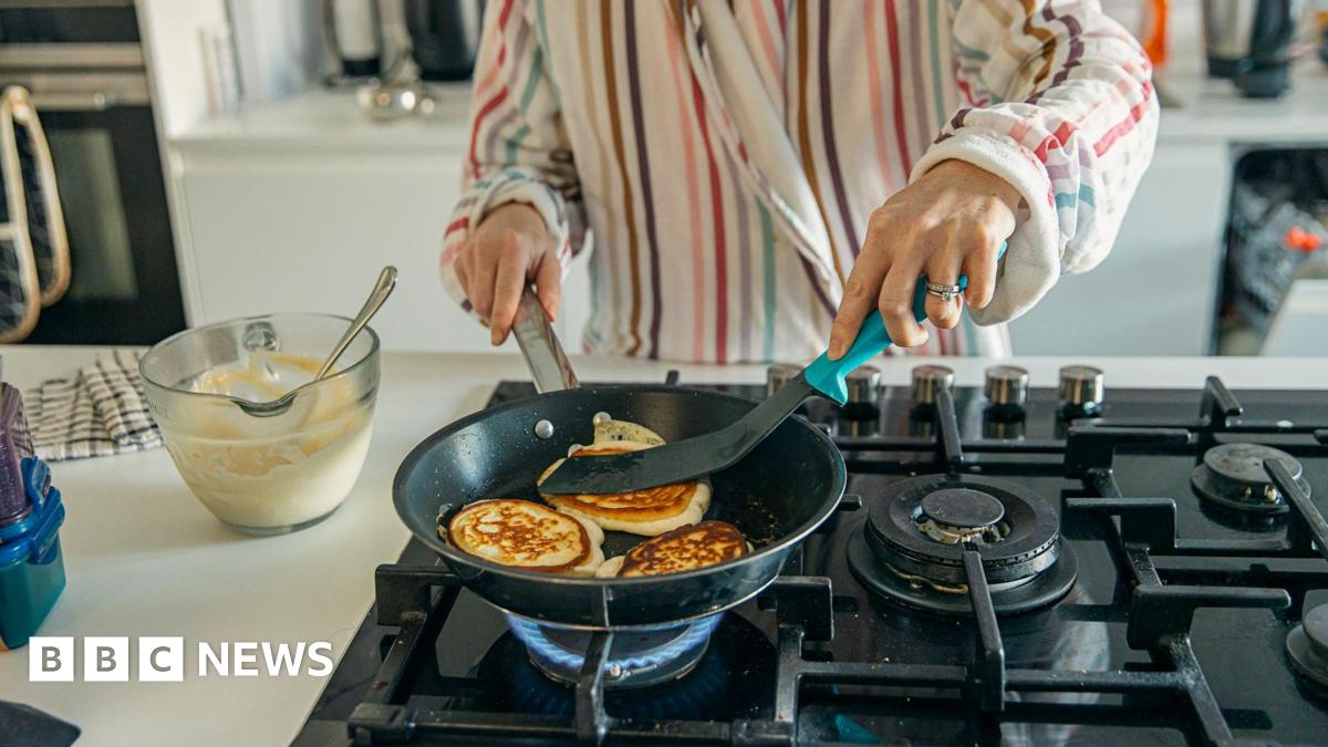 A woman dressed in striped pyjamas cooks pancakes over a gas stovetop. A glass bowl of pancake batter is on the bench beside her