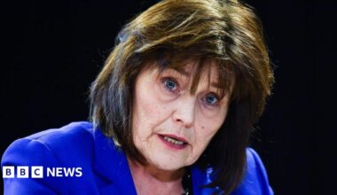 Jeane Freeman, a woman in her 60s with shoulder length dark brown hair, is wearing an electric blue jacket and is sitting talking against a black background.