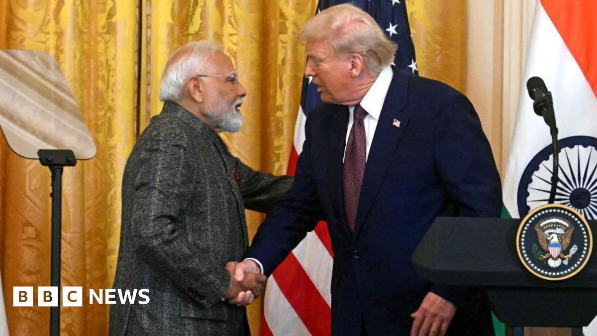 Indian Prime Minister Narendra Modi, wearing a grey suit, shakes hands with US President Donald Trump (in a blue suit, dark-coloured tie and white shirt) as they stand near a podium. Behind them, the Indian and US flags can be seen.