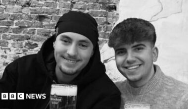 A black and white photo of Eddie and Jayden, smiling side by side. They are both holding pints or beer. Jayden has a hat on and has short stubble and is wearing a black hoodie. Eddie has short dark hair and is wearing a jumper.