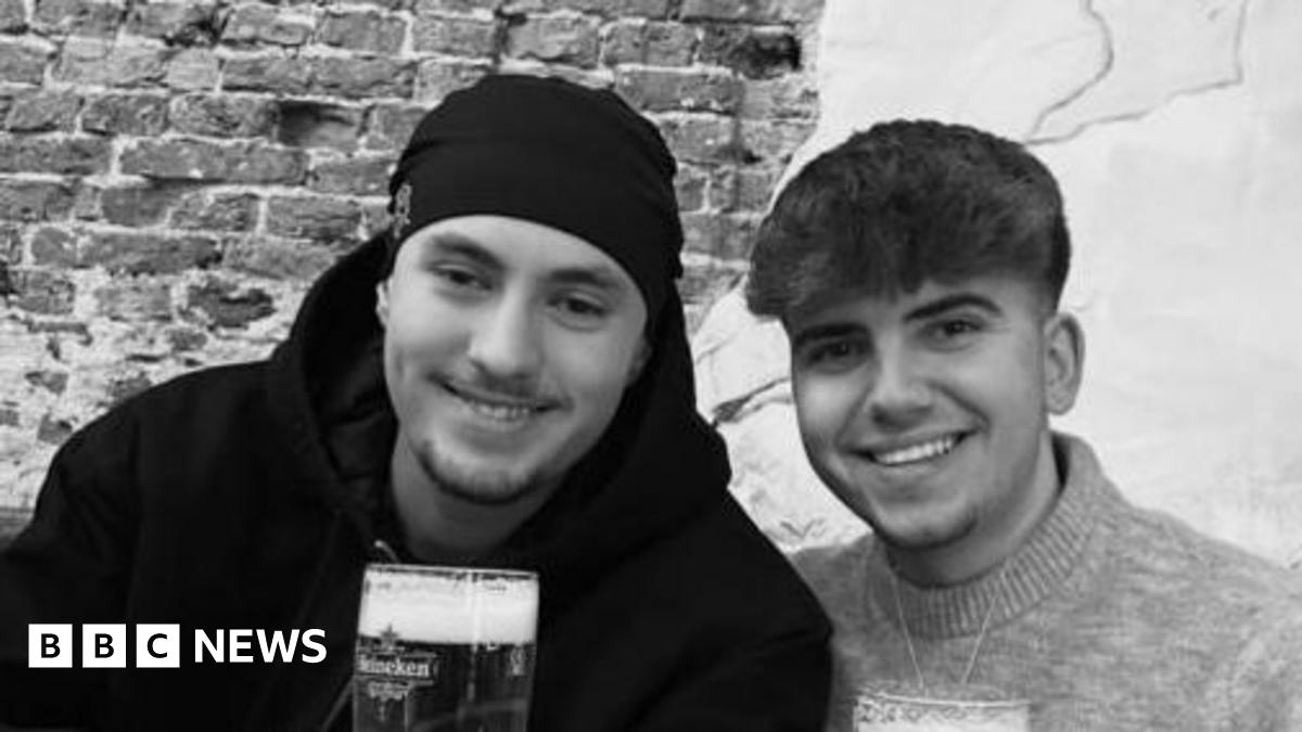 A black and white photo of Eddie and Jayden, smiling side by side. They are both holding pints or beer. Jayden has a hat on and has short stubble and is wearing a black hoodie. Eddie has short dark hair and is wearing a jumper.