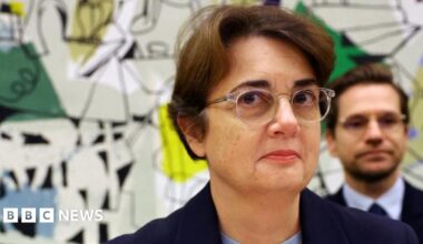 Head and shoulders picture of Laurence des Cars wearing glasses and standing in front of an artwork which is part visible behind her