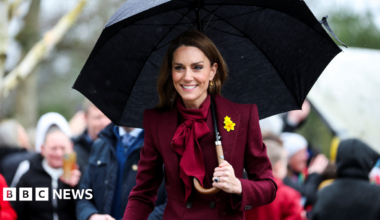 Catherine and William look to the right of the camera, both smiling. William has his mouth open and is holding his hand open in a greeting, he wears a daffodil on his lapel. Catherine wears a burgundy coat and her long brown hair is loose.
