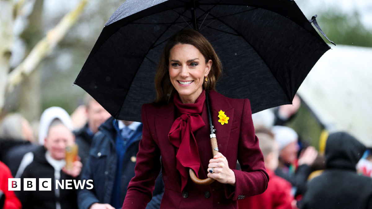Catherine and William look to the right of the camera, both smiling. William has his mouth open and is holding his hand open in a greeting, he wears a daffodil on his lapel. Catherine wears a burgundy coat and her long brown hair is loose.