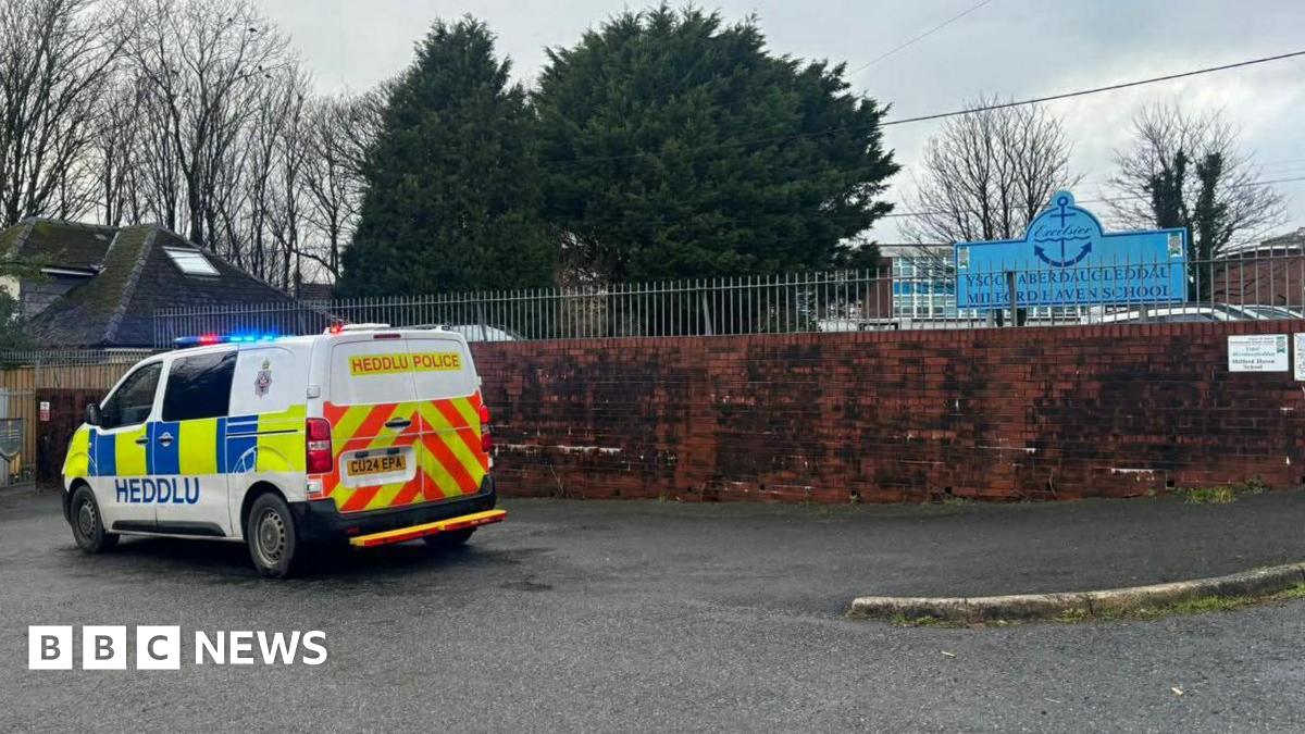 A police van and car are outside the entrance to the school. A sign with the school name can also be seen.