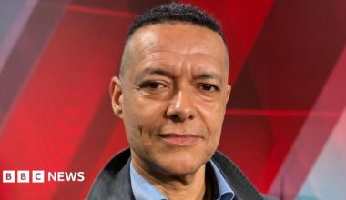 Clive Lewis stares at the camera. He is wearing an open necked blue shirt and a black jacket.