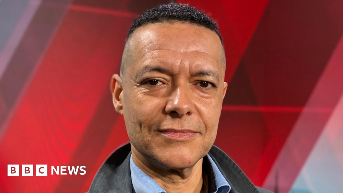 Clive Lewis stares at the camera. He is wearing an open necked blue shirt and a black jacket.