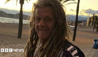 A smiling man with long dreadlocks. He is wearing a black t-shirt with white writing. He is stood on a beach and in the background is the sea and sand.