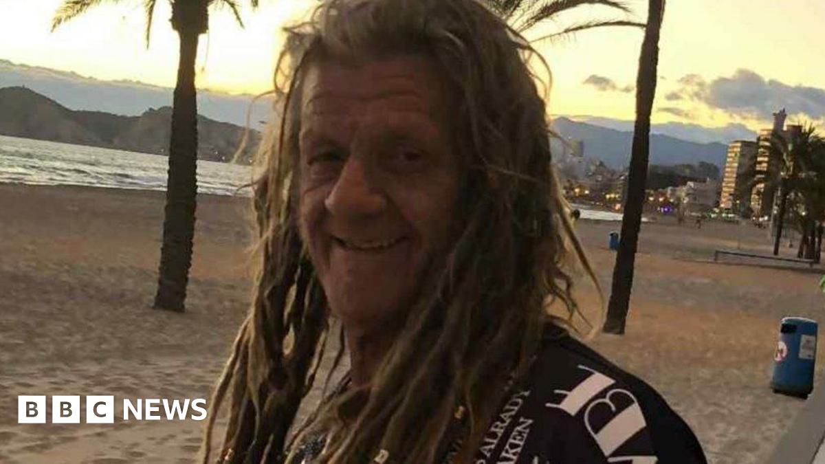 A smiling man with long dreadlocks. He is wearing a black t-shirt with white writing. He is stood on a beach and in the background is the sea and sand.