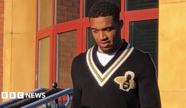 Jordon Ibe outside a building. He is wearing a black vest with a bee sewing on. He is looking down.