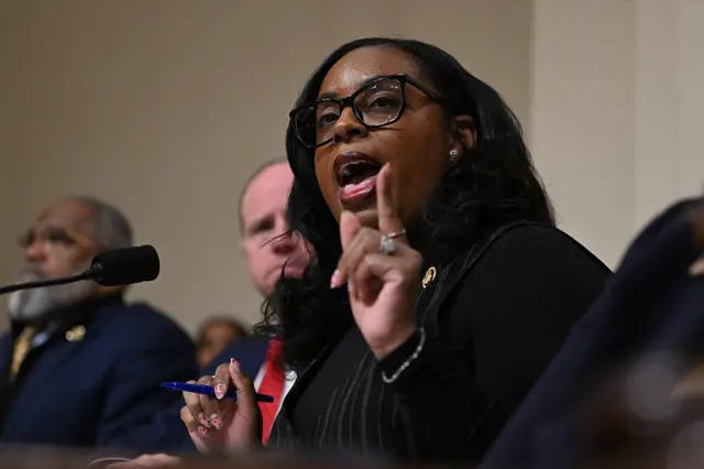 A woman with long hair, black-rimmed glasses and a dark suit gestures with one hand holds a blue pen with her other hand