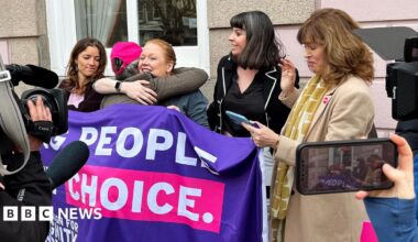 Five women celebrate with hugs next to a banner saying 'people, choice' with news cameras around them, outside on the street in Jersey on Thursday.