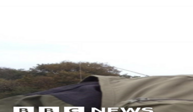 Alan Graham pictured holding a fish. He is wearing a khaki hat and fisherman's jacket. He has a grey beard and wears circular silver framed glasses