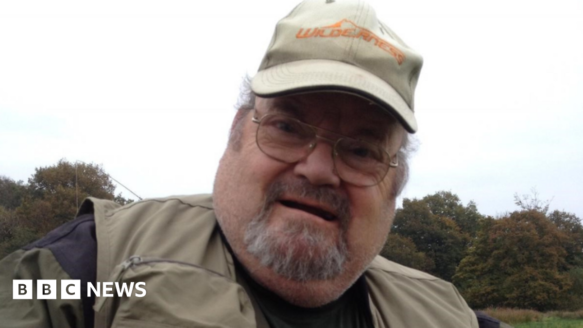Alan Graham pictured holding a fish. He is wearing a khaki hat and fisherman's jacket. He has a grey beard and wears circular silver framed glasses