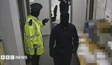 A gang member dressed as a police officer wearing a hi-vis jacket is standing with a gun in his hand. Two others, who are masked and wearing black are walking past him. There are notices on the wall in the depot.