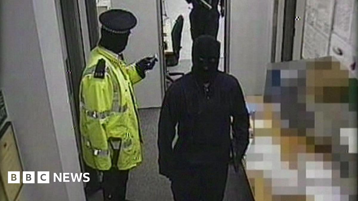 A gang member dressed as a police officer wearing a hi-vis jacket is standing with a gun in his hand. Two others, who are masked and wearing black are walking past him. There are notices on the wall in the depot.