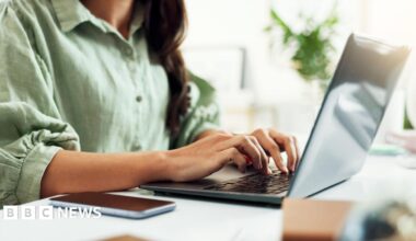 A woman with dark hair wearing a green button-up shirt with puffed three-quarter length sleeves. She is sitting at a white desk, typing on a grey laptop. In the background there is a plant out of focus.