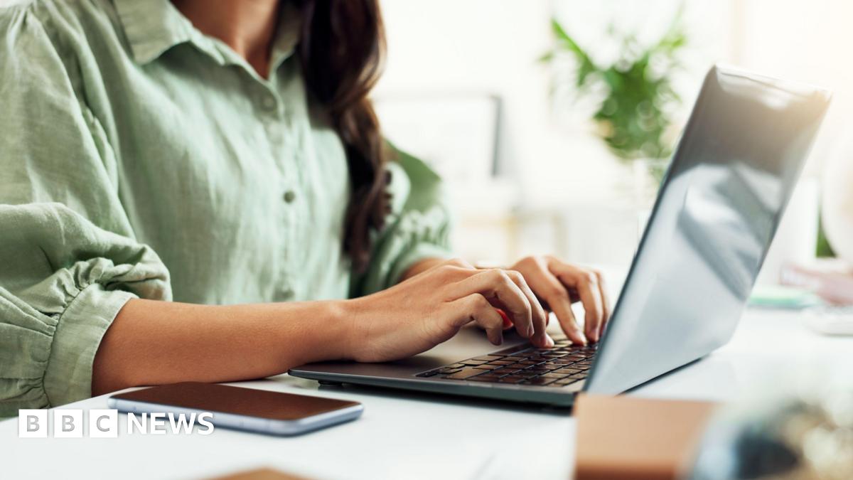 A woman with dark hair wearing a green button-up shirt with puffed three-quarter length sleeves. She is sitting at a white desk, typing on a grey laptop. In the background there is a plant out of focus.