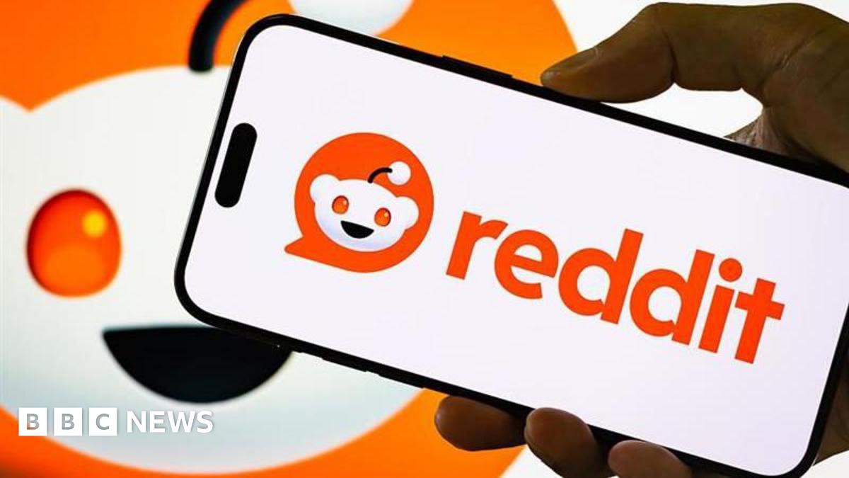 Reddit logo displayed on a smartphone held by hand, with a larger version of the company's logo behind it.