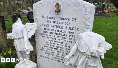 A white marble style headstone with a picture of a two-year-old boy and engraved with the name James Patrick Bulger. Either side are two cherub or angel statues with their heads smashed off.
