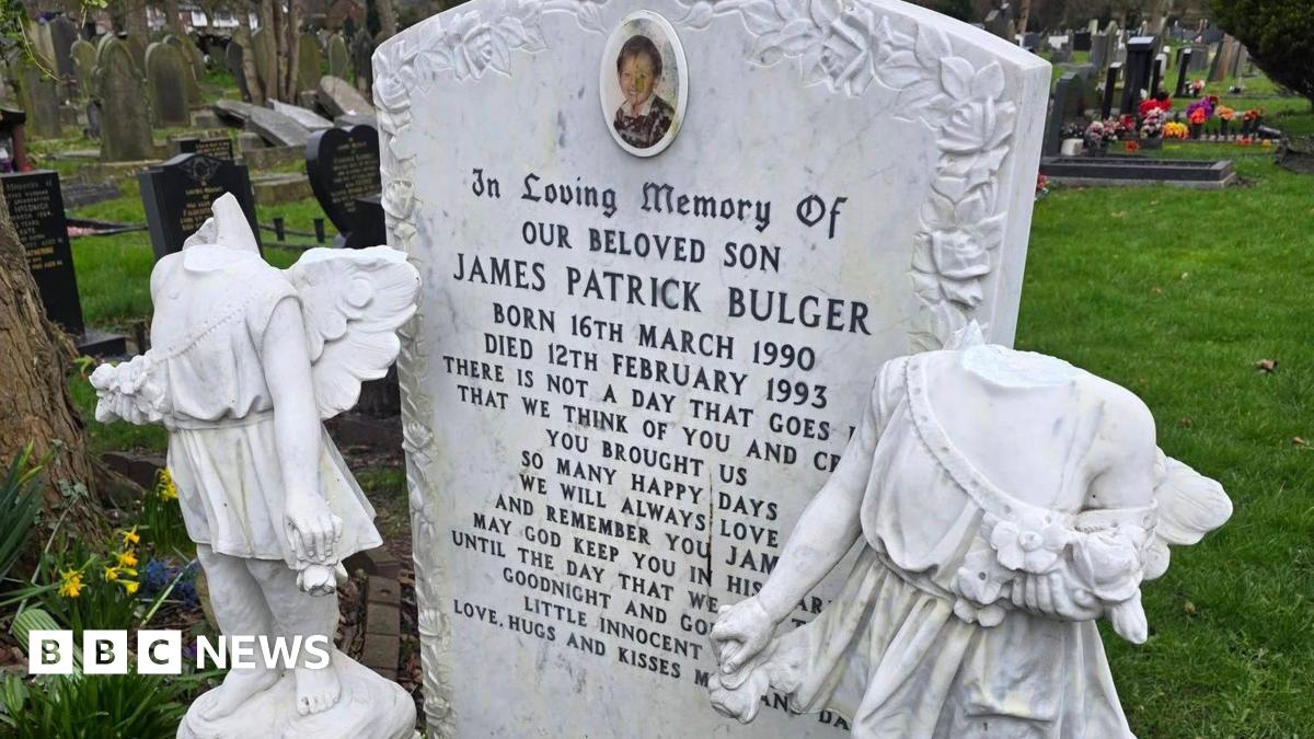 A white marble style headstone with a picture of a two-year-old boy and engraved with the name James Patrick Bulger. Either side are two cherub or angel statues with their heads smashed off.