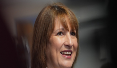 Chancellor Rachel Reeves smiles in a headshot. She wears a high-necked white shirt with a black jacket. Her light brown hair is work loose with a long fringe.