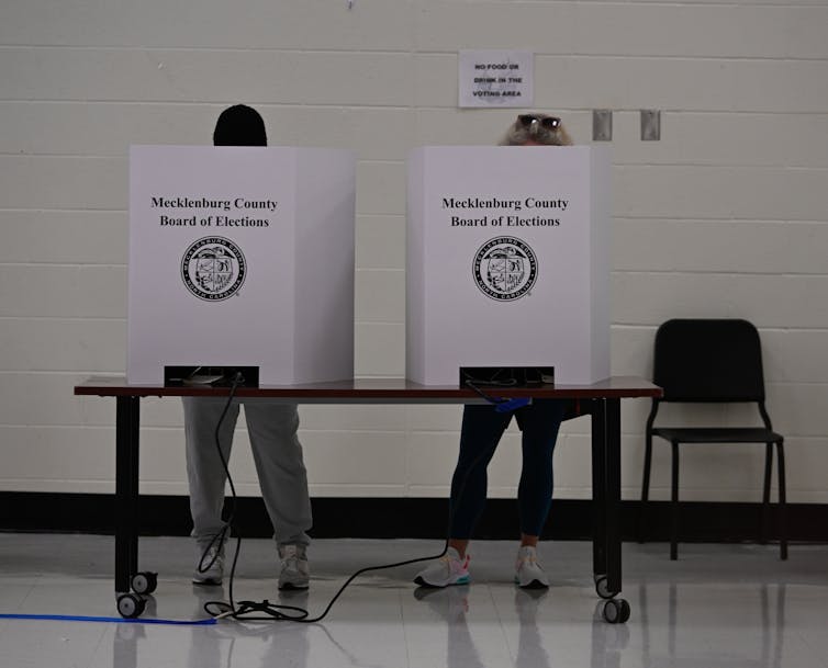 Two people stand behind large white voting machines that say 'Mecklenburg County Board of Elections' on them.