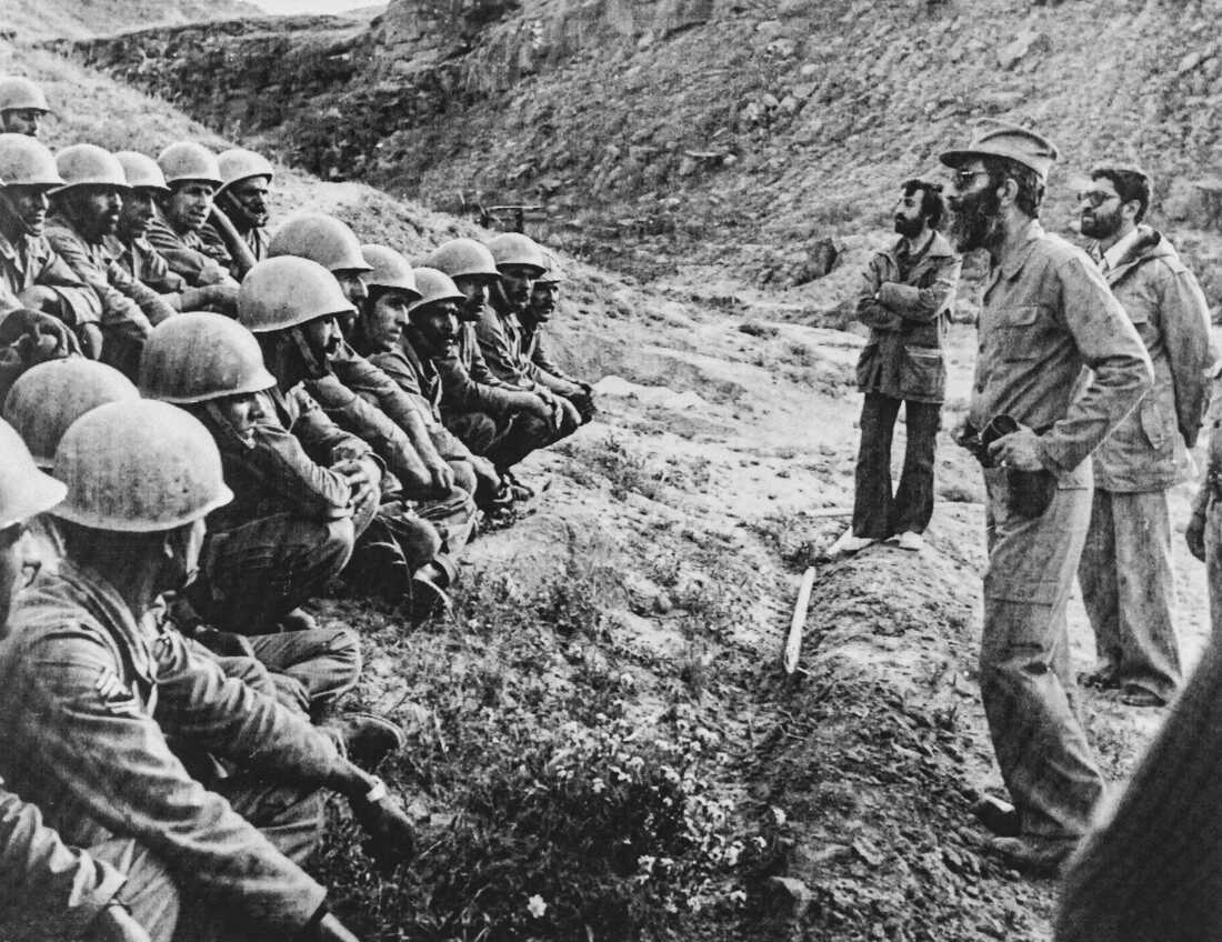 In this black-and-white photo from 1981, Ali Khamenei (on the right side of the frame) speaks to members of Iran's armed forces, who are wearing helmets and are seated or crouched in rows on the ground on the left side of the frame.
