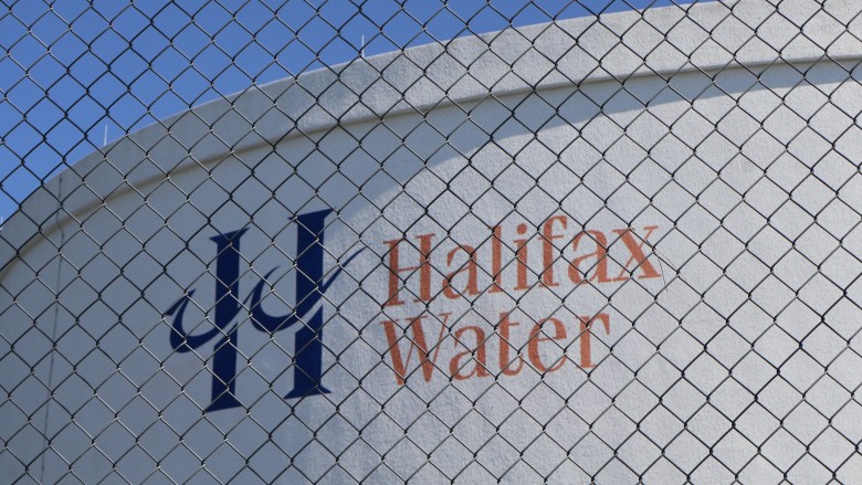 A white concrete water reservoir behind a black chain link fence. Halifax Water is painted in orange on the side with a large letter "H" painted in blue.