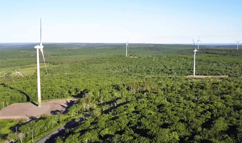 An overhead view on a sunny day shows at least five large white industrial windmills set in a lush green landscape with a road running through the middle of the land.