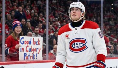 Patrik Laine, wearing a white Montreal Canadiens away jersey and helmet, looks up while on the ice. Behind the glass boards, a young female fan wearing a Canadiens toque holds a handmade sign with blue and red lettering that reads, "Good luck in Calgary Patrik!".
