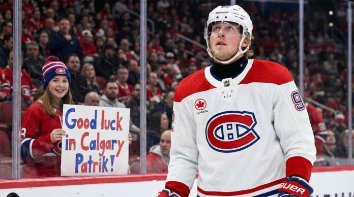 Patrik Laine, wearing a white Montreal Canadiens away jersey and helmet, looks up while on the ice. Behind the glass boards, a young female fan wearing a Canadiens toque holds a handmade sign with blue and red lettering that reads, "Good luck in Calgary Patrik!".