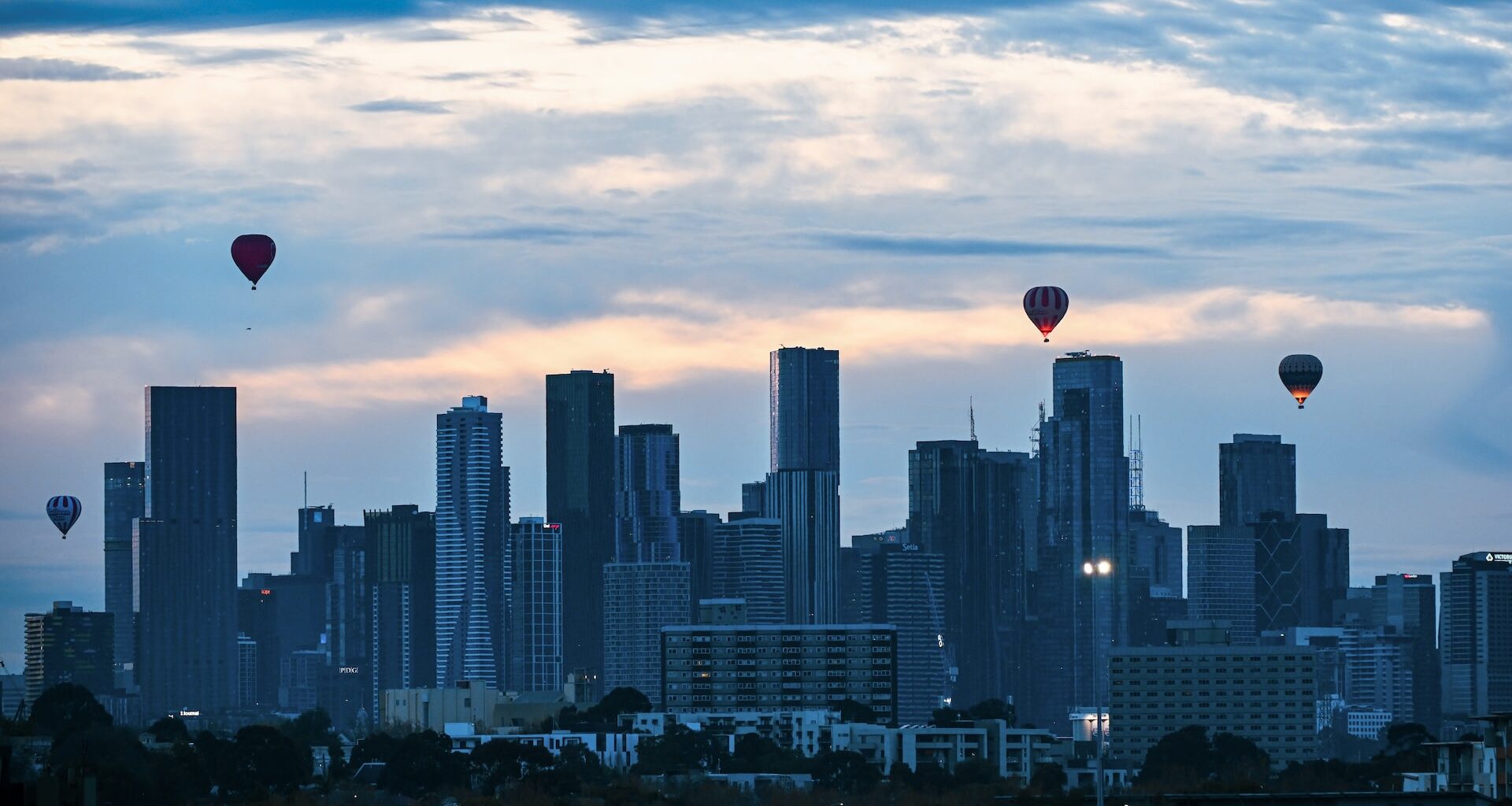 A city skyline at dawn with several hot air balloons floating in the sky above tall buildings.