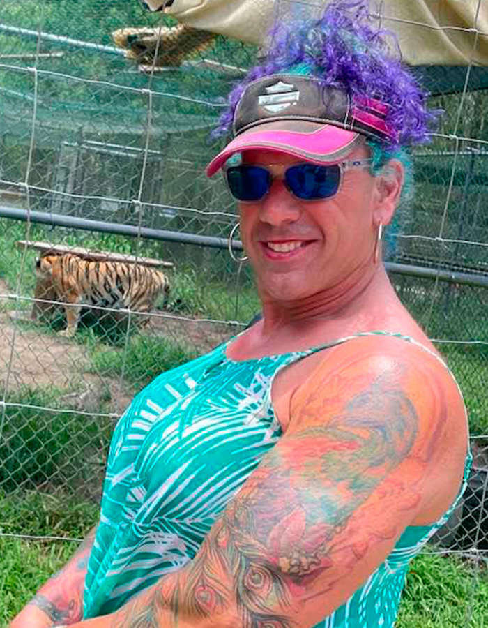 Person with colorful hair and tattoos smiling outdoors near fenced area with a tiger, related to ice rink attacker family discovery. Person with colorful hair and tattoos smiling outdoors near fenced area with a tiger, related to ice rink attacker family discovery.