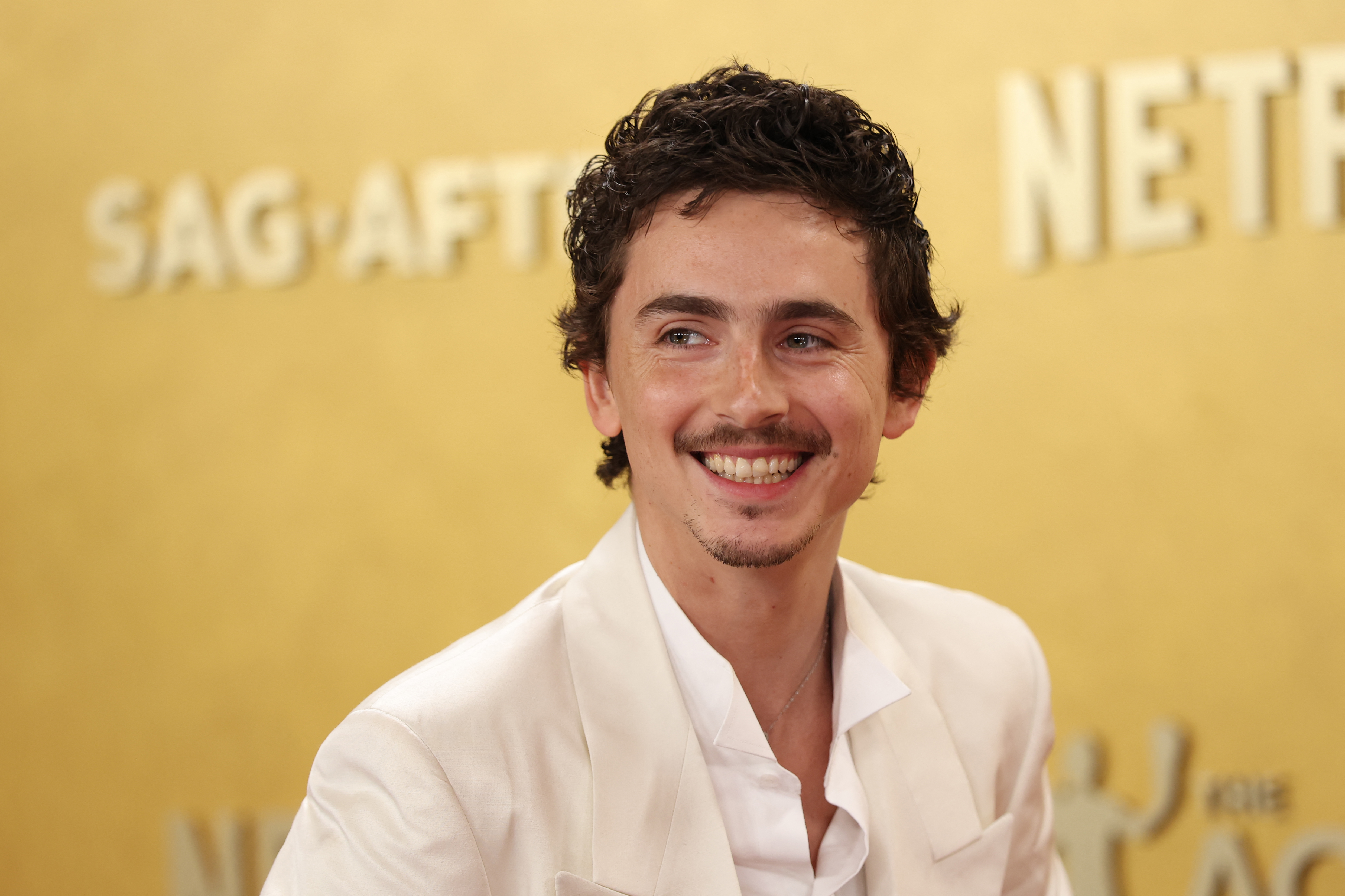 Timothee Chalamet smiling in a white suit jacket and unbuttoned white shirt, on a gold background.