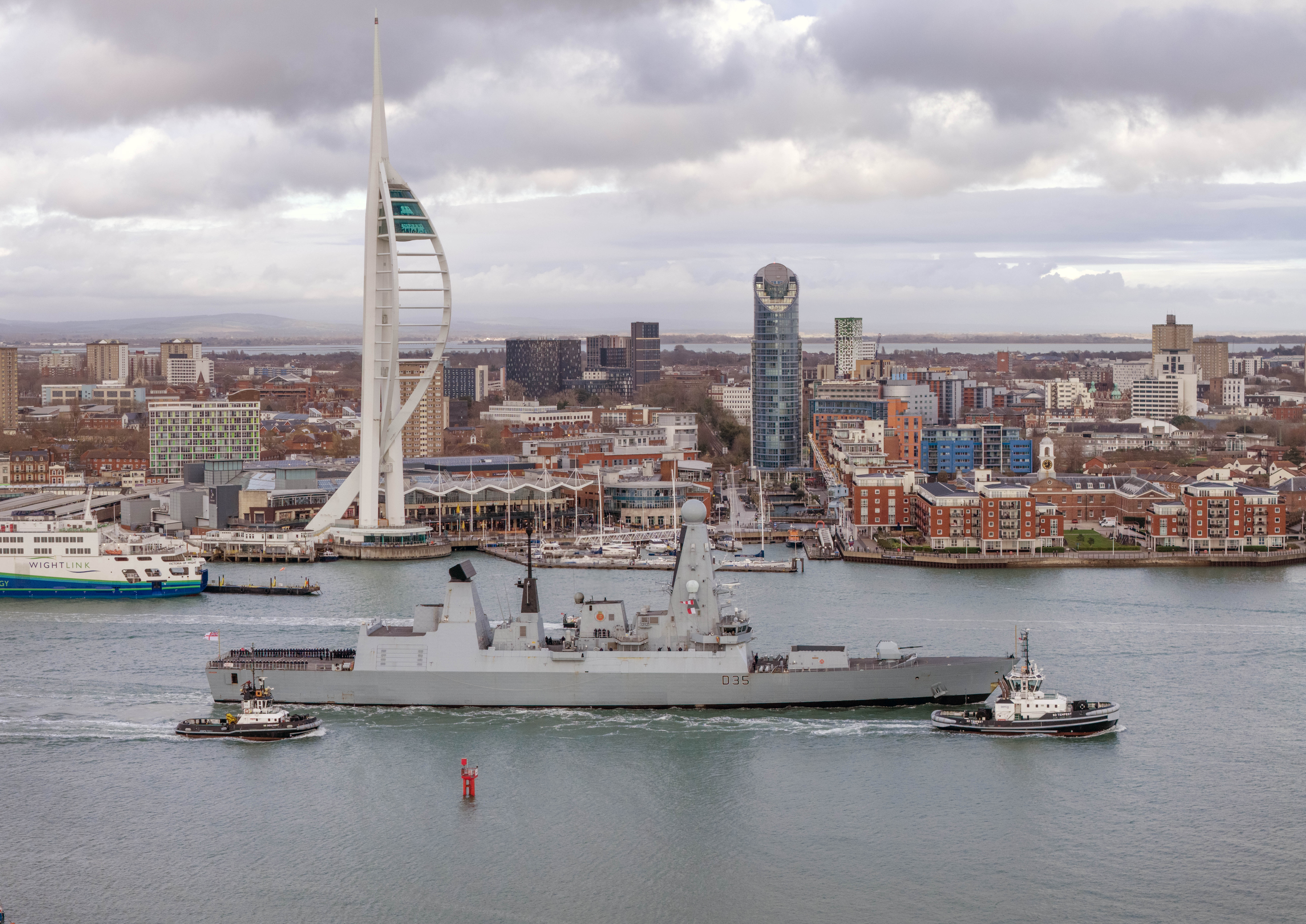 HMS Dragon, a grey warship, departs Portsmouth Harbour, flanked by two tugboats, with the Spinnaker Tower and city skyline in the background.